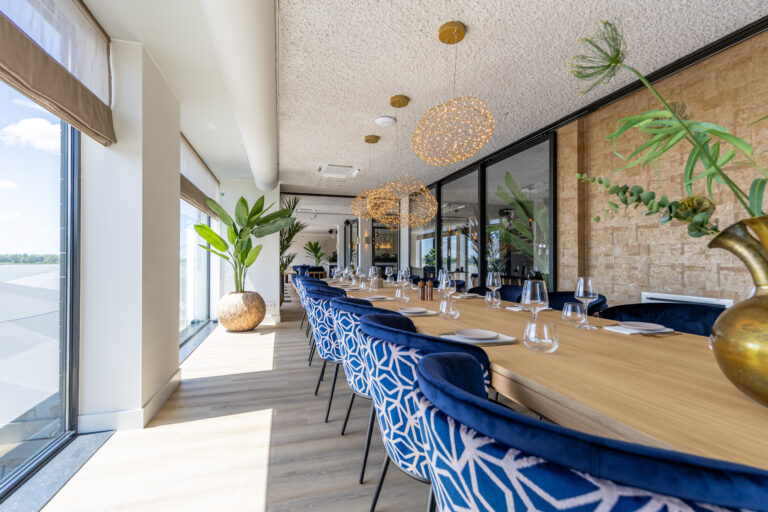Lange houten eettafel met gedekte plaatsen en blauwe fluwelen stoelen in een moderne zaal met ramen en gouden hanglampen.