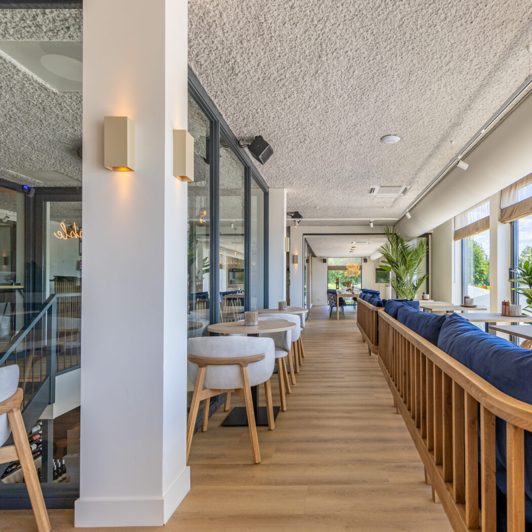 Licht, modern restaurantinterieur met lange houten vloer, blauwe zitbanken, lichte stoelen en grote ramen. Een wijnkelder is zichtbaar.