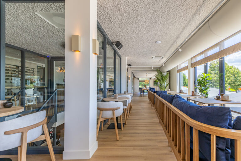 Licht, modern restaurantinterieur met lange houten vloer, blauwe zitbanken, lichte stoelen en grote ramen. Een wijnkelder is zichtbaar.