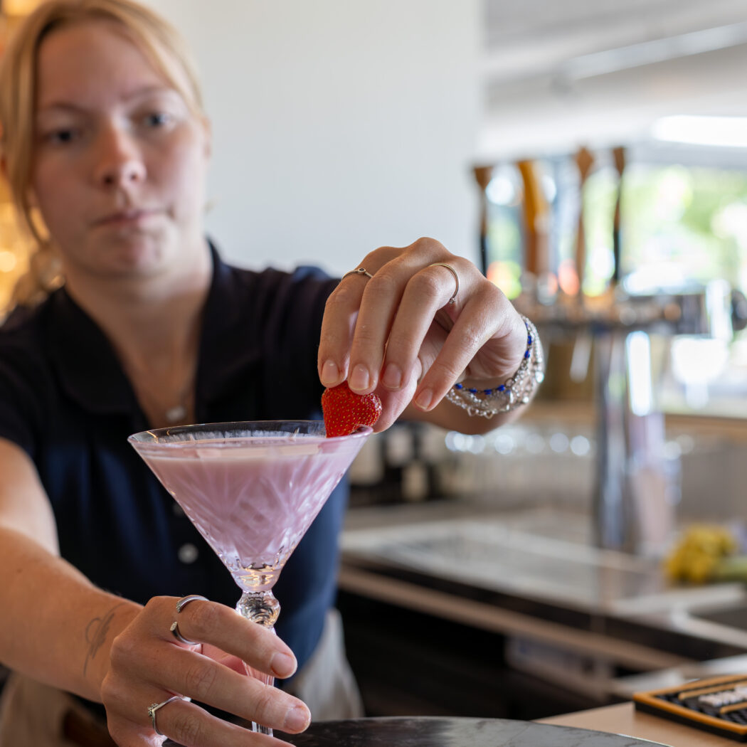 Een barvrouw garneert een roze cocktail in een coupeglas met een aardbei.