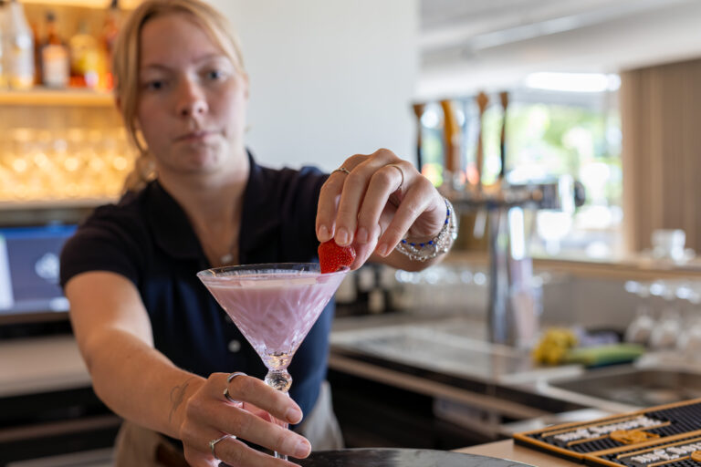 Een barvrouw garneert een roze cocktail in een coupeglas met een aardbei.