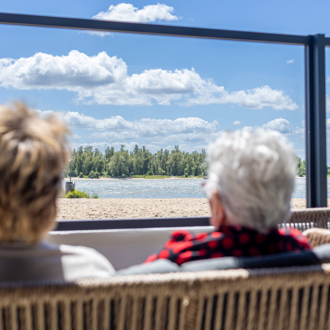 Twee mensen zitten op een rieten bank achter een glazen balustrade met uitzicht op zandstrand, water en bomen.