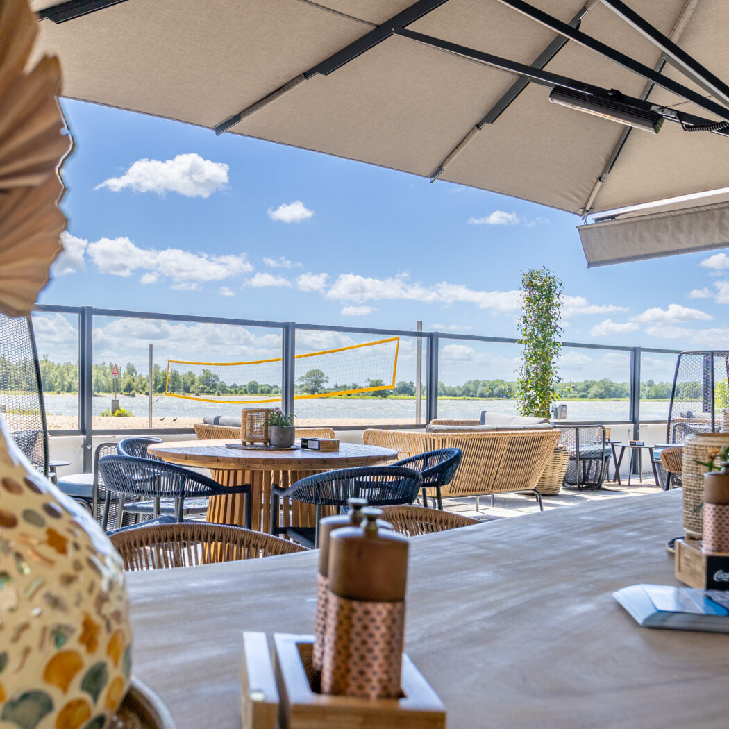 Zonnig terras met beige parasols en comfortabele zitjes, uitkijkend op een strand met beachvolleybalnet en water.