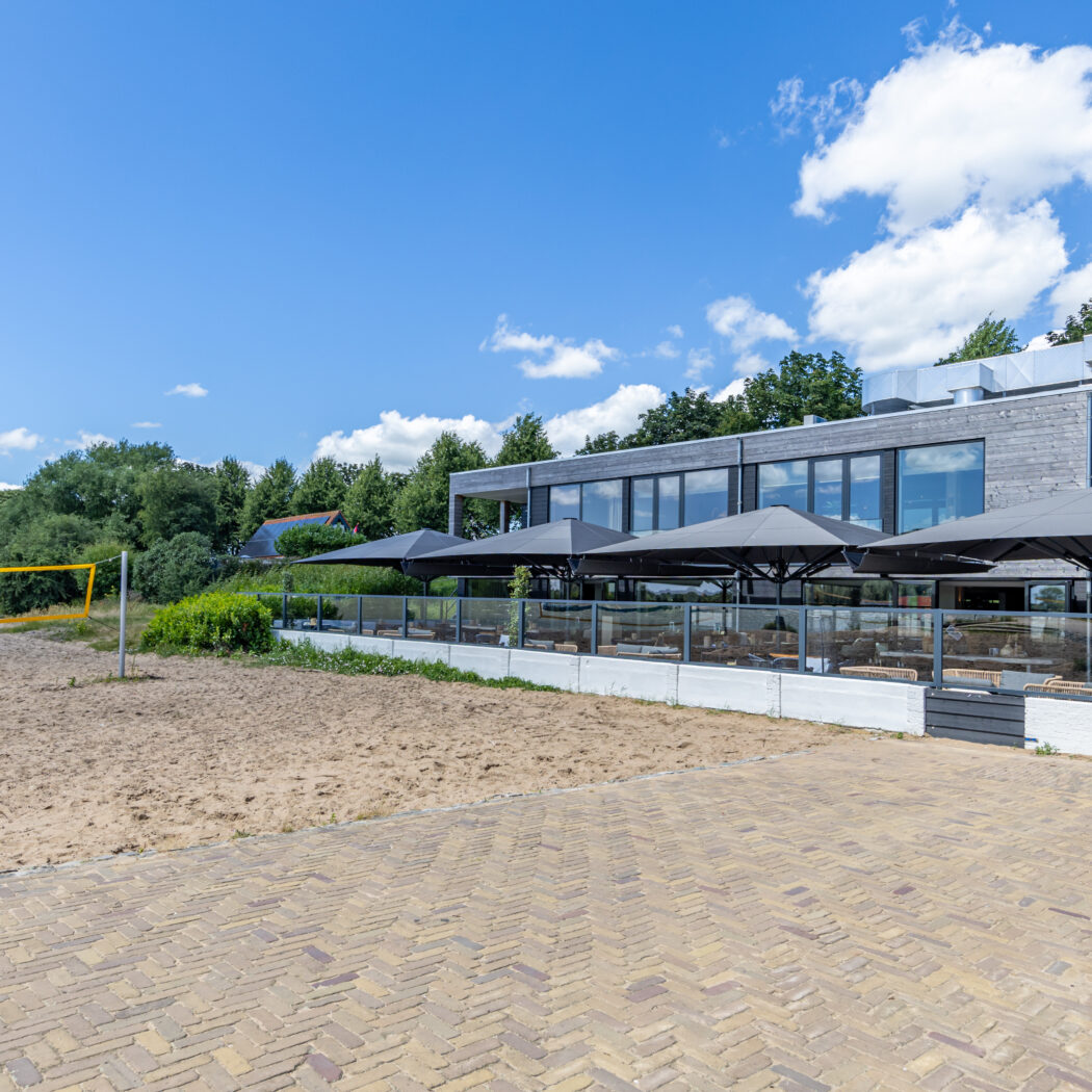 Modern gebouw met houten gevel, terras met grijze parasols en een zandvolleybalveld.