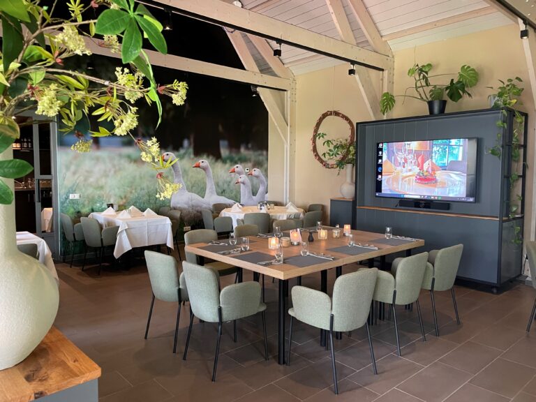 Ruim restaurant of evenementenlocatie met gedekte tafels, een muurschildering van ganzen en een tv-scherm.