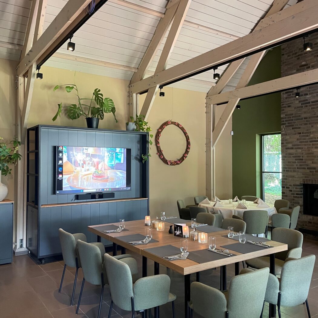 Sfeervolle eetzaal met een grote vierkante houten tafel, groene stoelen en tv-scherm onder een balkenplafond.