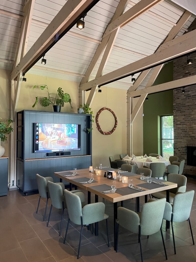 Sfeervolle eetzaal met een grote vierkante houten tafel, groene stoelen en tv-scherm onder een balkenplafond.