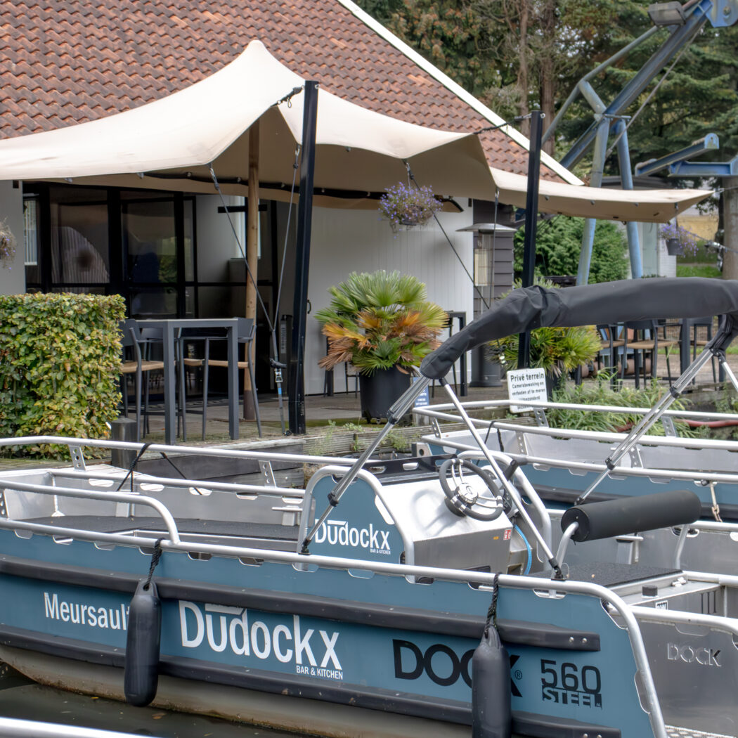 Blauwe Dudockx sloep aangemeerd bij restaurant Dudockx BAR & KITCHEN met terras onder een beige overkapping.