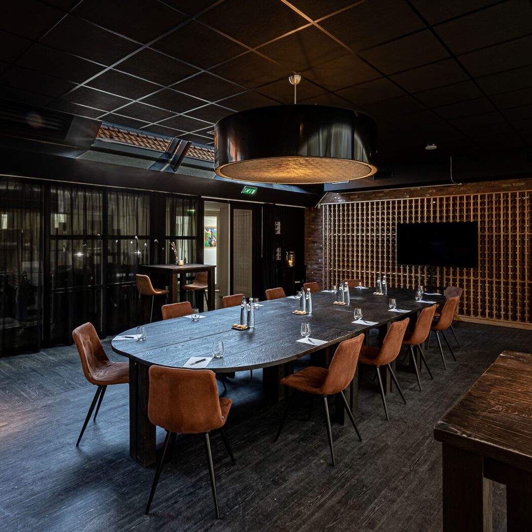 Luxe ovale vergadertafel in donkere ruimte met cognackleurige stoelen, wijnrek en scherm.