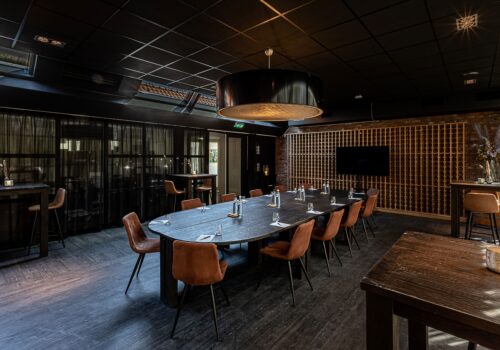 Luxe ovale vergadertafel in donkere ruimte met cognackleurige stoelen, wijnrek en scherm.