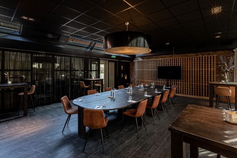 Luxe ovale vergadertafel in donkere ruimte met cognackleurige stoelen, wijnrek en scherm.